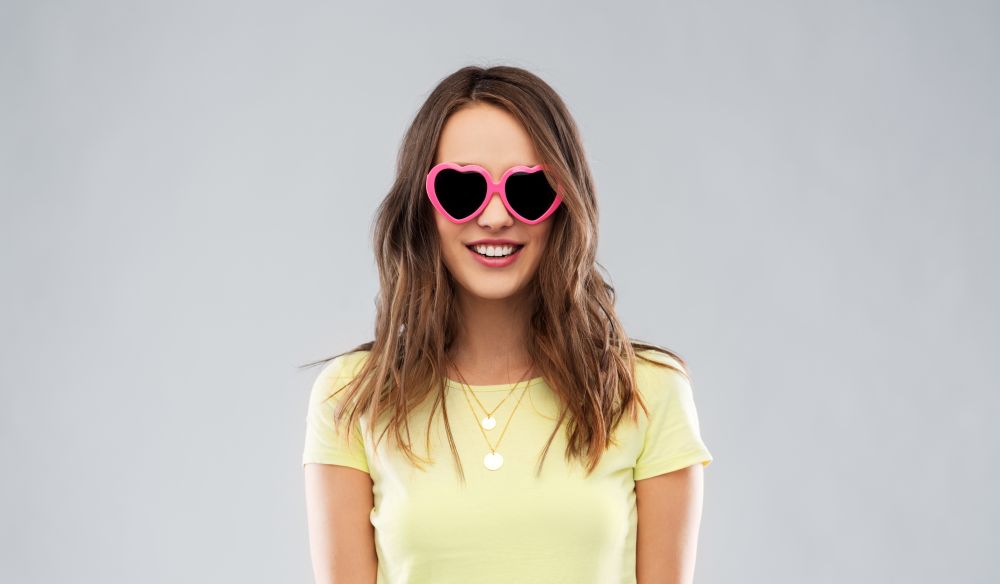 summer, valentine&rsquo;s day and people concept - smiling young woman or teenage girl in yellow t-shirt and heart-shaped sunglasses over grey background. teenage girl in heart-shaped sunglasses