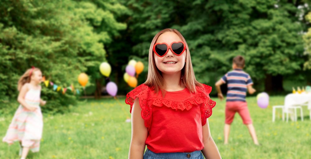 childhood, valentine&rsquo;s day and summer concept - smiling preteen girl with heart shaped sunglasses over birthday party at park. girl in heart shaped sunglasses at birthday party