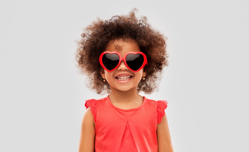 childhood, valentine&rsquo;s day and summer concept - happy little african american girl in heart shaped sunglasses over grey background. african ameican girl in heart shaped sunglasses