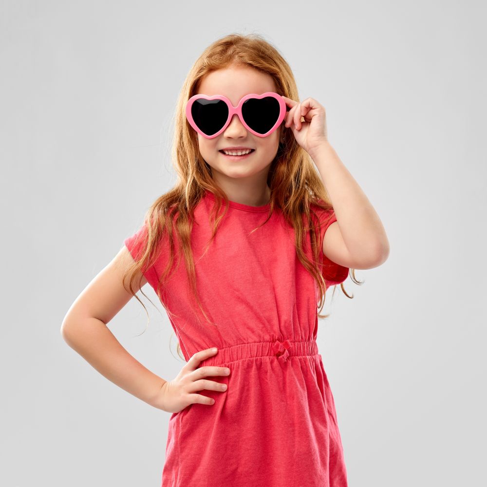 childhood, valentine&rsquo;s day and summer concept - smiling red haired preteen girl in heart shaped sunglasses over grey background. smiling red haired girl in heart shaped sunglasses