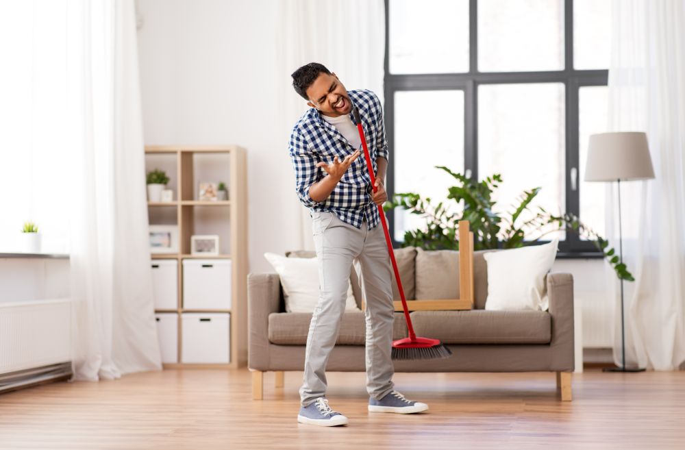 cleaning, housework and housekeeping concept - indian man with broom sweeping floor and singing at home. man with broom cleaning and singing at home