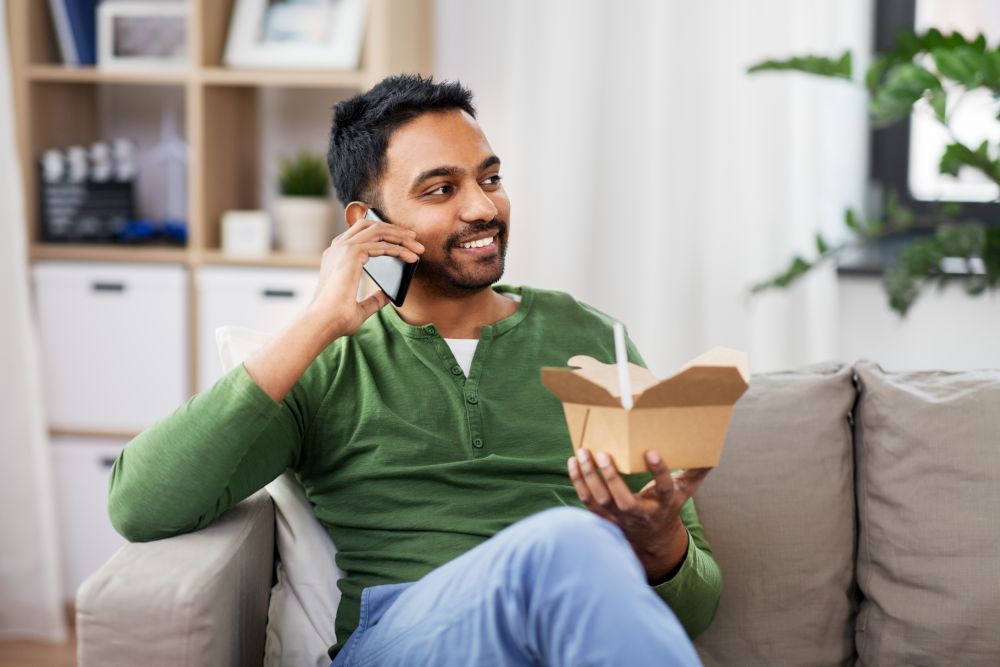communication, leisure and people concept - smiling indian man calling on smartphone and eating takeaway food at home. smiling indian man eating takeaway food at home