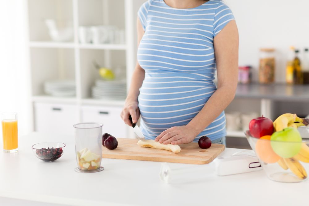 cooking, pregnancy and healthy eating concept - pregnant woman with kitchen knife chopping fruits on wooden cutting board at home. pregnant woman chopping fruits at home kitchen