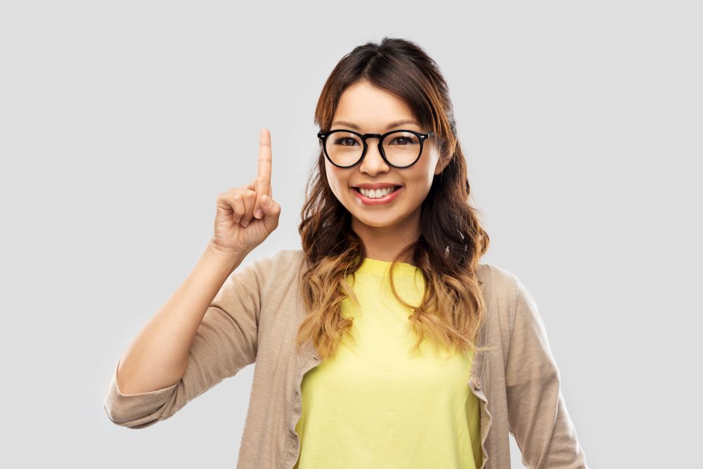 people, education and school concept - happy asian woman in glasses or student pointing finger up over grey background. asian woman in glasses or student with finger up