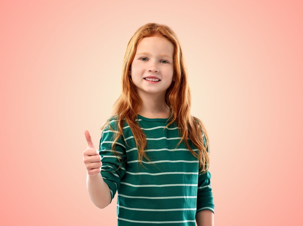 childhood, fashion and people concept - smiling red haired girl in green striped shirt showing thumbs up over pink living coral background. smiling red haired girl showing thumbs up