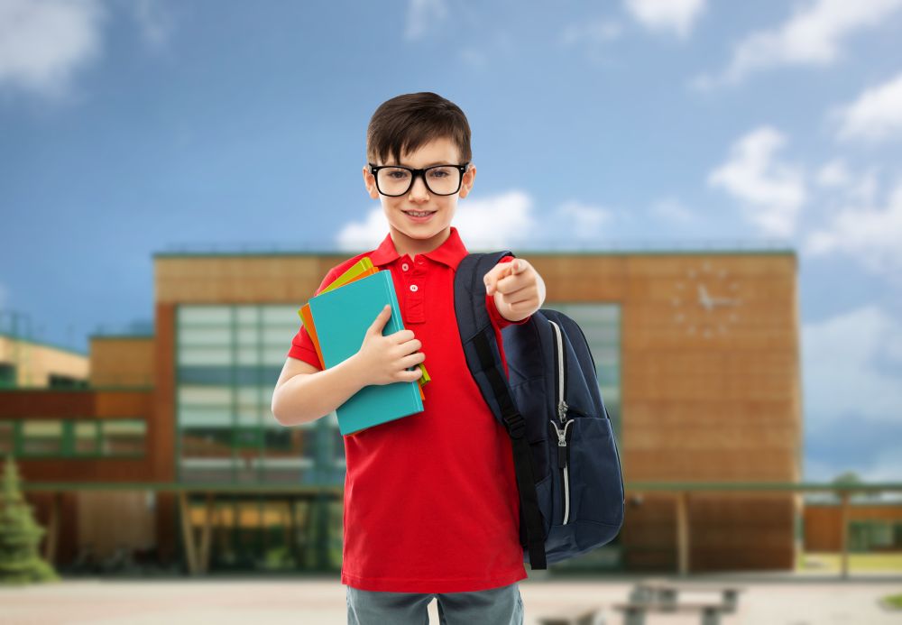 education and people concept - portrait of smiling little student boy in red polo t-shirt in glasses with books and bag over school background. schoolboy with books and bag over school