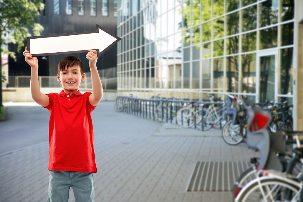 direction, childhood and people concept - portrait of nice little boy in red polo t-shirt holding big white rightwards thick arrow over school yard background. boy holding big white rightwards thick arrow