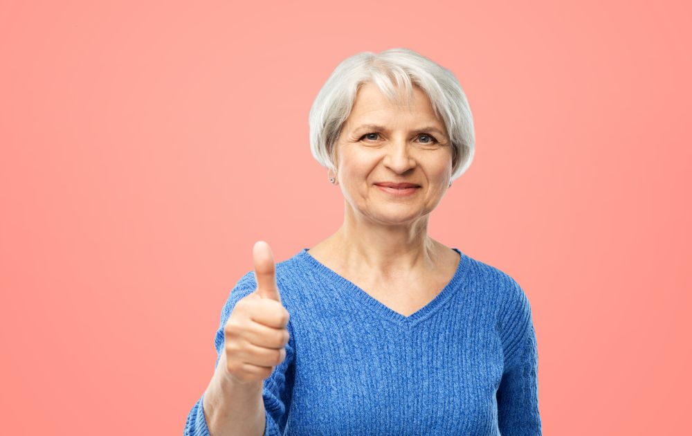 gesture and old people concept - portrait of smiling senior woman in blue sweater showing thumbs up over pink or living coral background. smiling senior woman r showing thumbs up over pink