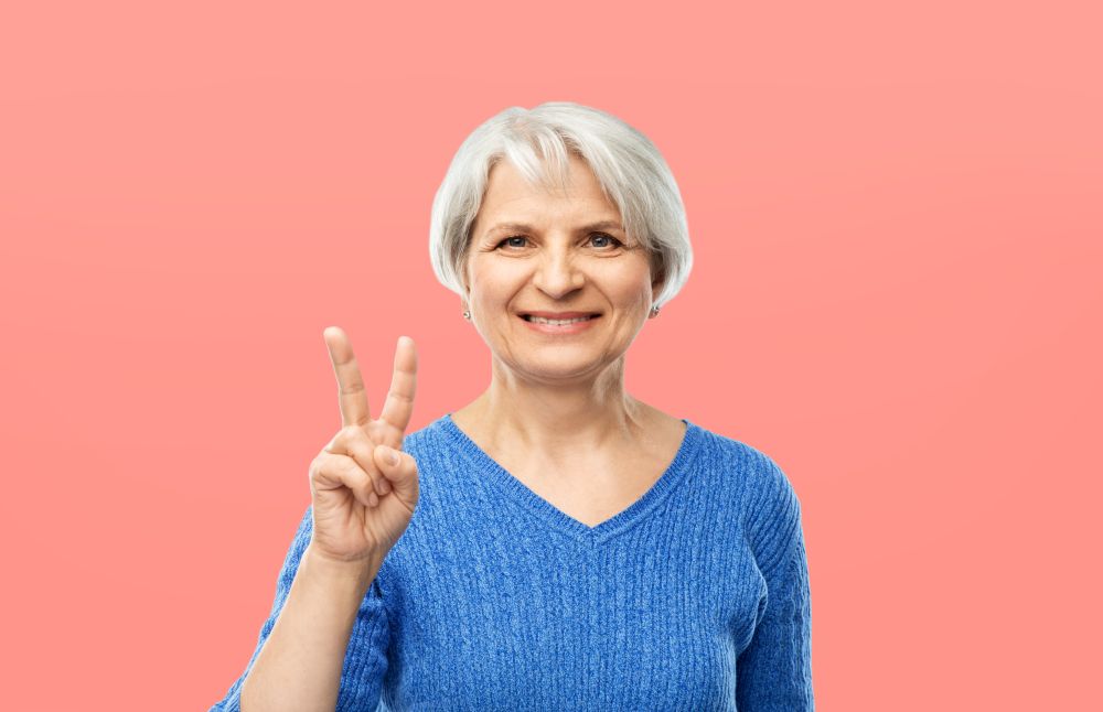 gesture and old people concept - portrait of smiling senior woman in blue sweater showing peace over pink or living coral background. portrait of smiling senior woman showing peace