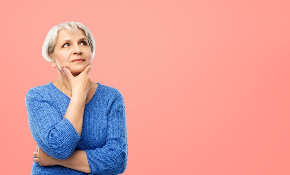 old people and decision making concept - portrait of senior woman in blue sweater thinking over pink or living coral background. portrait of senior woman in blue sweater thinking