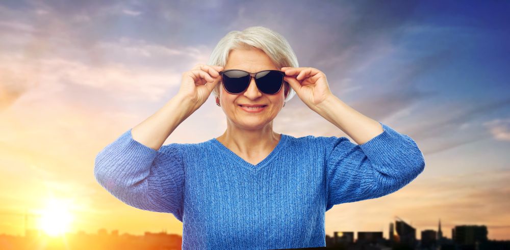 valentine&rsquo;s day, summer and old people concept - portrait of smiling senior woman in red black sunglasses over sunset in tallinn city background. senior woman in sunglasses over sunset in city