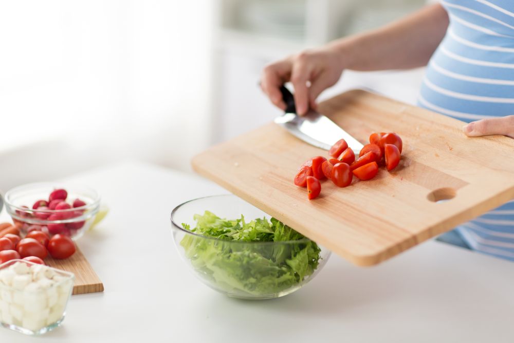 pregnancy, cooking food and healthy eating concept - close up of pregnant woman with kitchen knife adding chopped cherry tomato from wooden cutting board to salad bowl at home. close up of pregnant woman cooking food at home