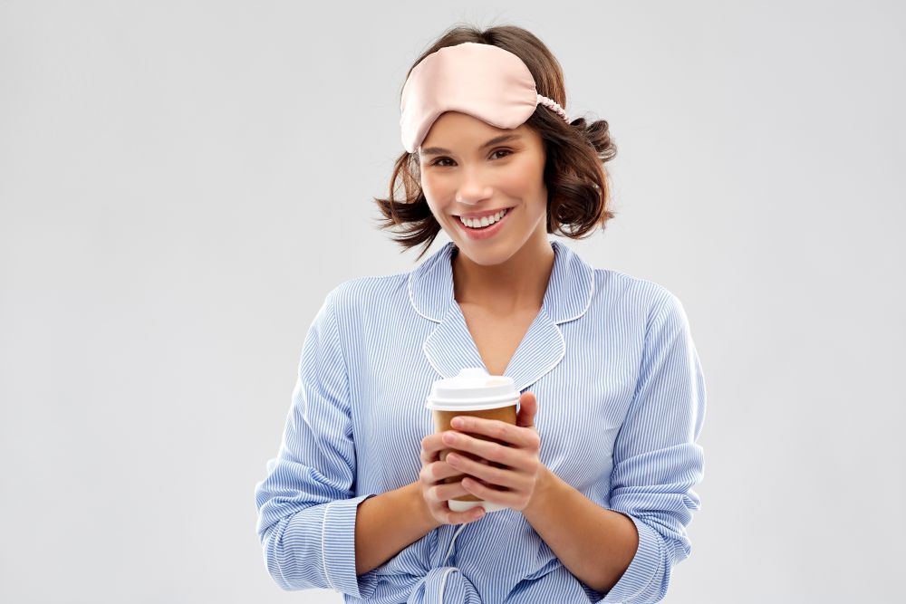 people and bedtime concept - happy young woman in pajama and eye sleeping mask with takeaway paper cup of coffee over grey background. woman in pajama and sleeping mask with coffee cup