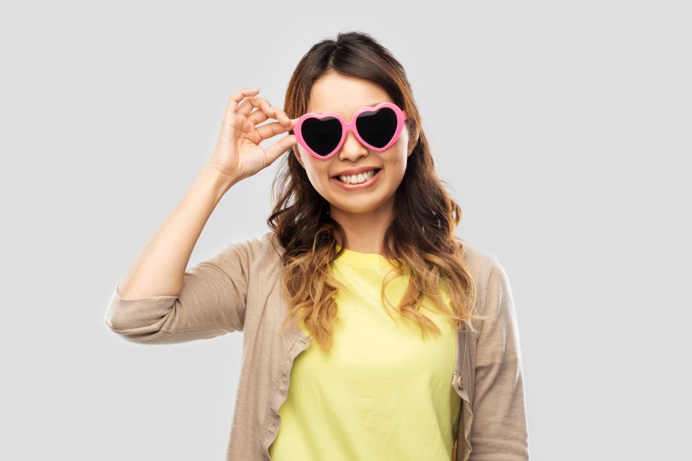 summer, valentine&rsquo;s day and eyewear concept - happy asian young woman in pink heart-shaped sunglasses over grey background. asian woman in heart-shaped sunglasses