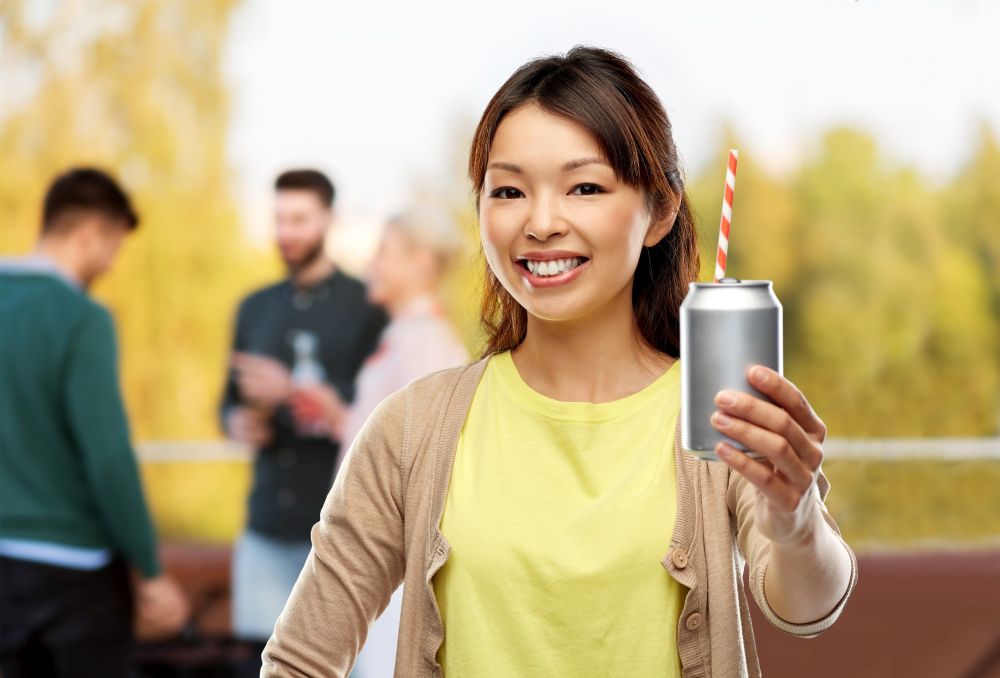 drinks and people concept - happy young asian woman drinking soda from can with paper straw over rooftop party background. asian woman with can drink over rooftop party