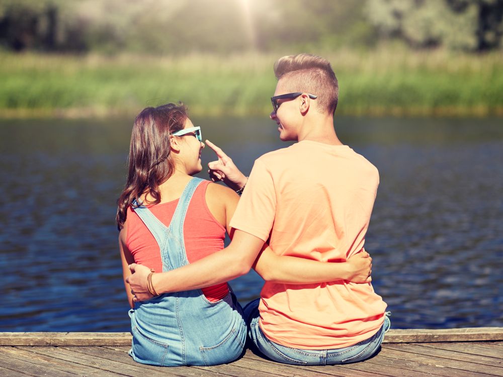 vacation, love and people concept - happy teenage couple sitting and hugging on river berth at summer. happy teenage couple hugging on river summer berth