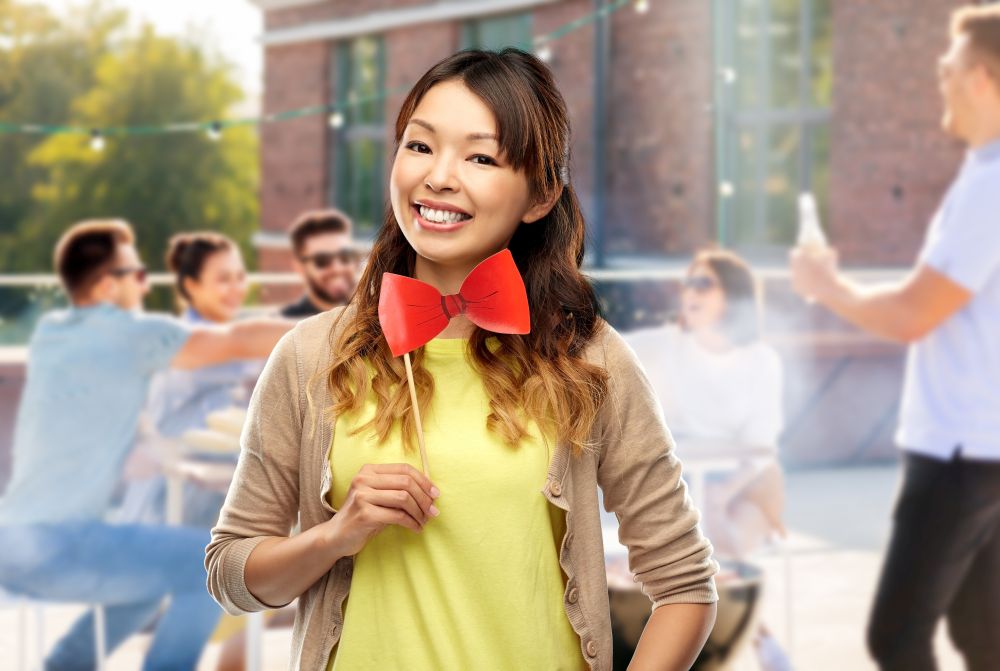 leisure and people concept - happy asian young woman with big red bow tie over rooftop party background. happy asian woman with bow tie over rooftop party