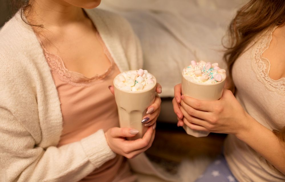 drinks, hygge and pajama party concept - close up of two women or female friends drinking hot chocolate with marshmallow at home. two women drinking hot chocolate with marshmallow