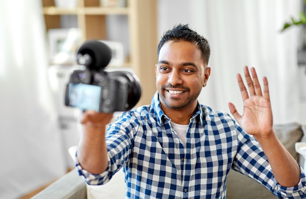 blogging, videoblog and people concept - smiling indian male video blogger with camera videoblogging and waving hand at home. male video blogger with camera blogging at home