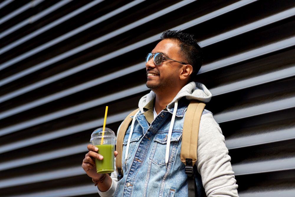 travel, tourism and lifestyle concept - smiling indian man with backpack drinking smoothie from plastic cup with straw on city street. man with backpack drinking smoothie on street
