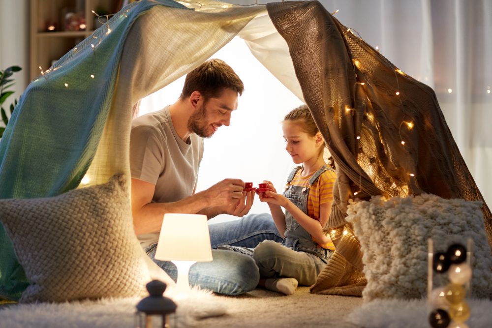 family, hygge and people concept - happy father and little daughter playing tea party in kids tent at night at home. family playing tea party in kids tent at home