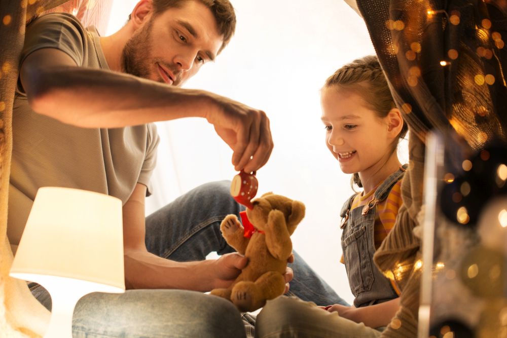 family, hygge and people concept - happy father and little daughter playing with teddy bear and toy tea pot in kids tent at night at home. happy family playing with toy in kids tent at home