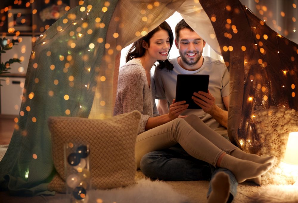 leisure, hygge, technology and people concept - happy couple with tablet pc computer in kids tent at home. happy couple with tablet pc in kids tent at home