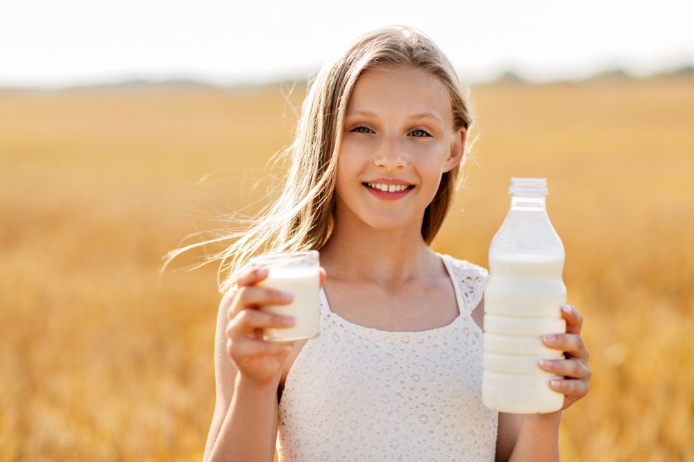 nature, healthy eating and organic concept - smiling young girl holding bottle and glass of milk on cereal field in summer. girl with bottle and glass of milk on cereal field