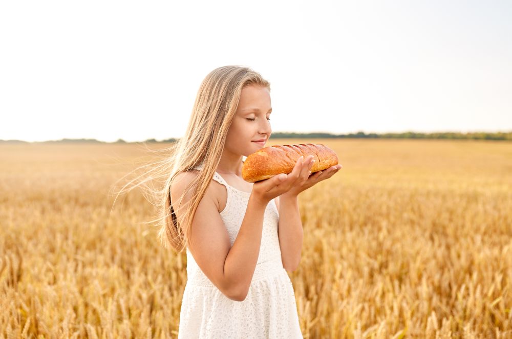 nature, healthy eating and harvesting concept - smiling young girl smelling loaf of white bread on cereal field in summer. girl smelling loaf of white bread on cereal field