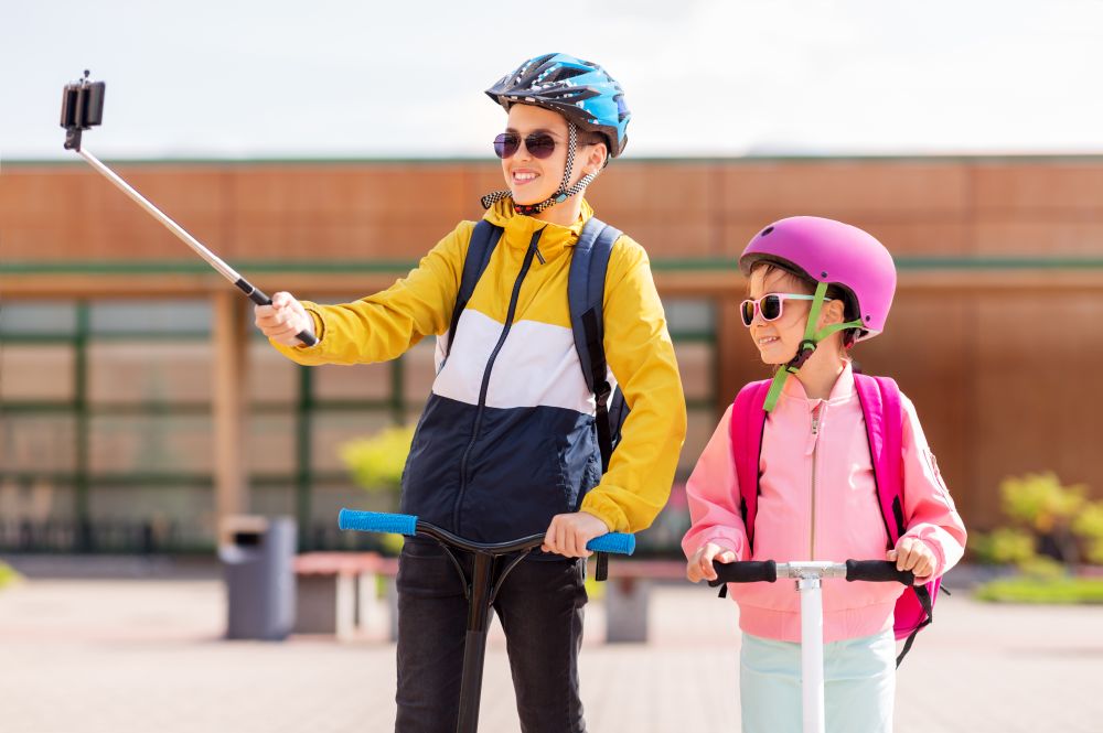 education, childhood and people concept - happy school children in helmets with backpacks riding scooters and taking picture by smartphone on selfie stick outdoors. happy school kids with scooters taking selfie