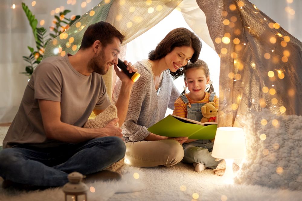 family, hygge and people concept - happy mother, father and little daughter reading book with torch light in kids tent at night at home. happy family reading book in kids tent at home