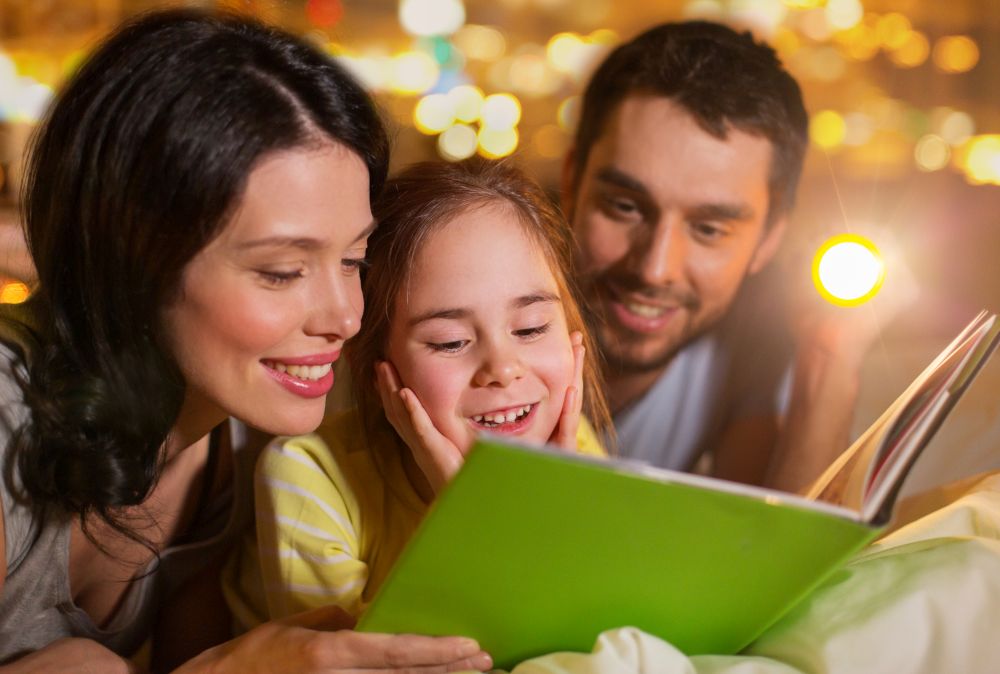 people and family concept - happy mother, father and little daughter reading book with torch light in bed at night at home. happy family reading book in bed at night at home