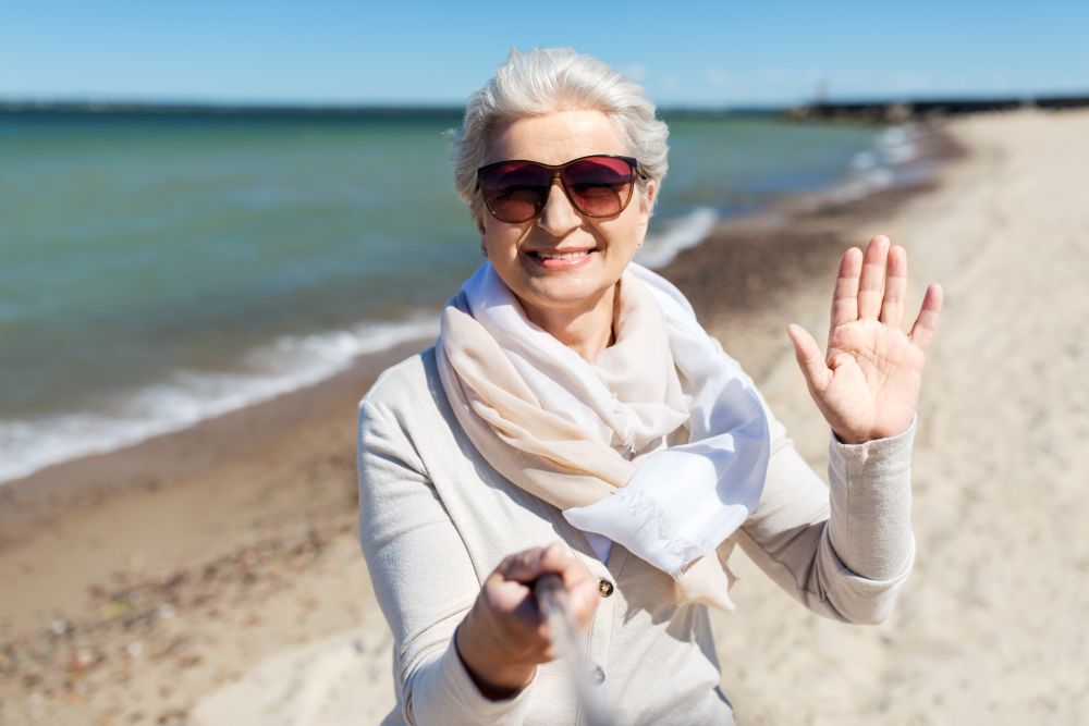 old people and leisure concept - happy smiling senior woman in sunglasses taking picture by selfie stick and waving hand on beach in estonia. old woman taking selfie and waving hand on beach