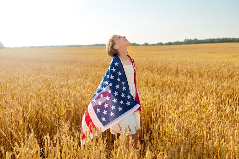 patriotism, independence day and country concept - happy smiling young girl wrapped into national american flag on cereal field looking up. happy girl in american flag on cereal field