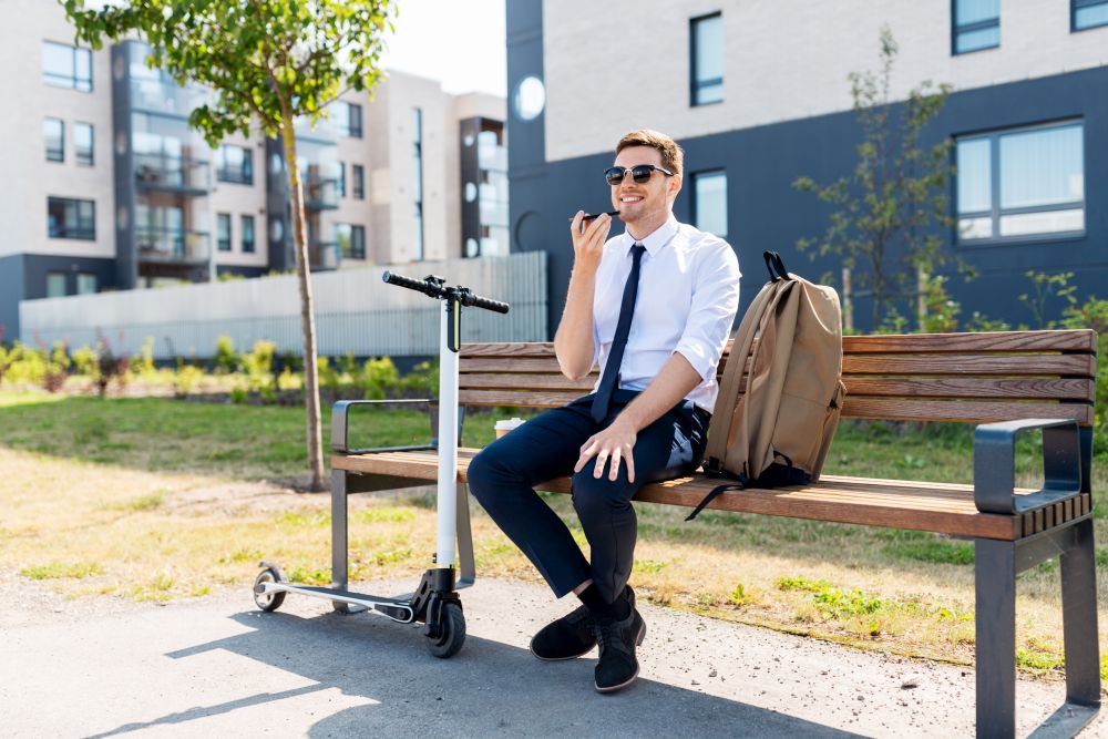 business and people and concept - young smiling businessman with backpack and scooter sitting on street bench and using voice command recorder on smartphone in city. businessman recording voice message by smartphone