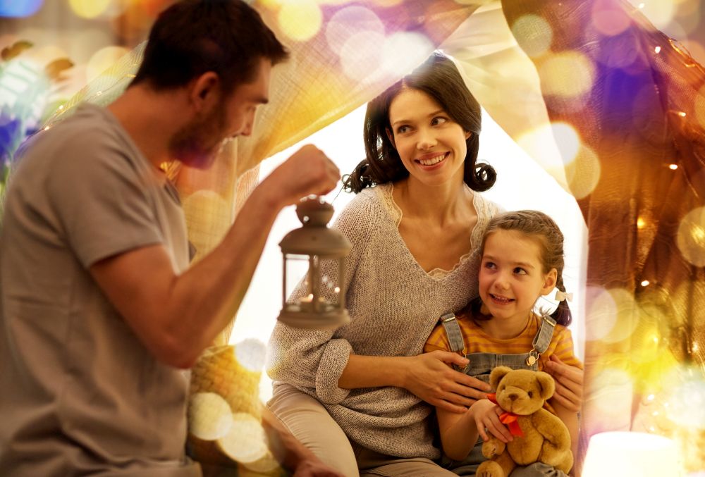 family, hygge and people concept - happy mother, father with lantern and little daughter with teddy bear playing in kids tent at night at home. happy family playing in kids tent at night at home