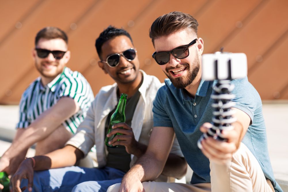 leisure, technology and people concept - happy male friends taking selfie by smartphone on tripod and drinking beer on street in summer. men drinking beer and taking selfie by smartphone