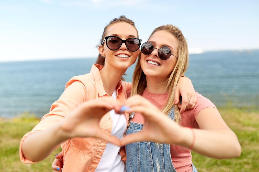 leisure and friendship concept - happy smiling teenage girls or best friends in sunglasses at seaside in summer hugging and making hand heart gesture. teenage girls or best friends at seaside in summer