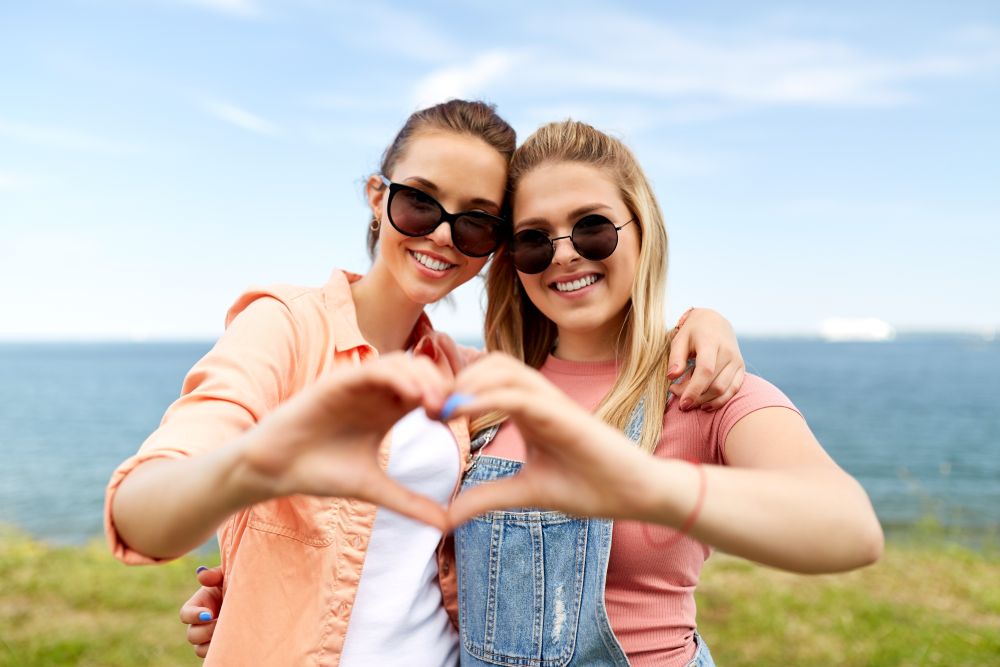 leisure and friendship concept - happy smiling teenage girls or best friends in sunglasses at seaside in summer hugging and making hand heart gesture. teenage girls or best friends at seaside in summer