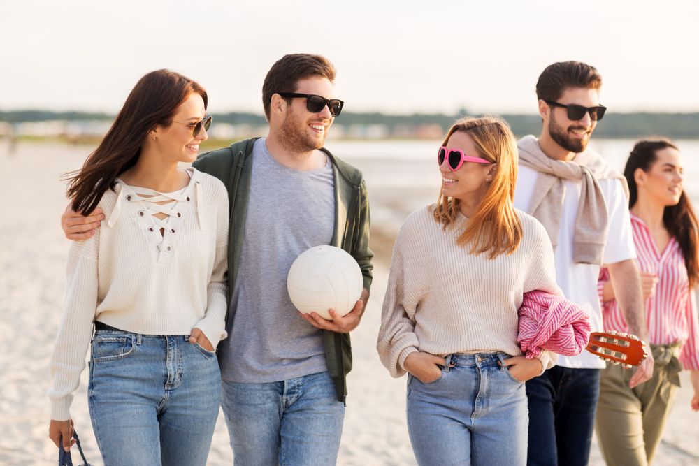 friendship, leisure and people concept - group of happy friends with ball, guitar, bag and blanket walking along beach in summer. happy friends walking along summer beach