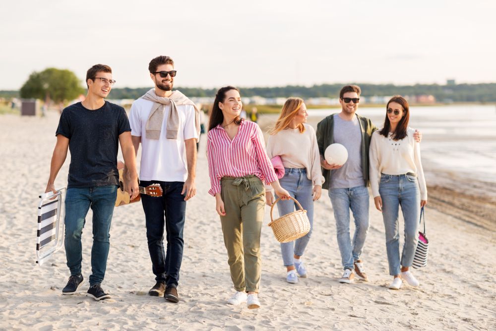 friendship, leisure and people concept - group of happy friends with guitar, chair and picnic basket walking along beach in summer. happy friends walking along summer beach