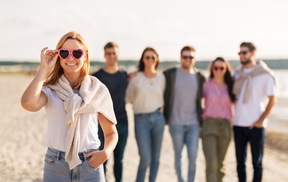 friendship, valentine&rsquo;s day and people concept - happy woman in heart-shaped sunglasses with group of friends on beach in summer. happy woman with friends on beach in summer