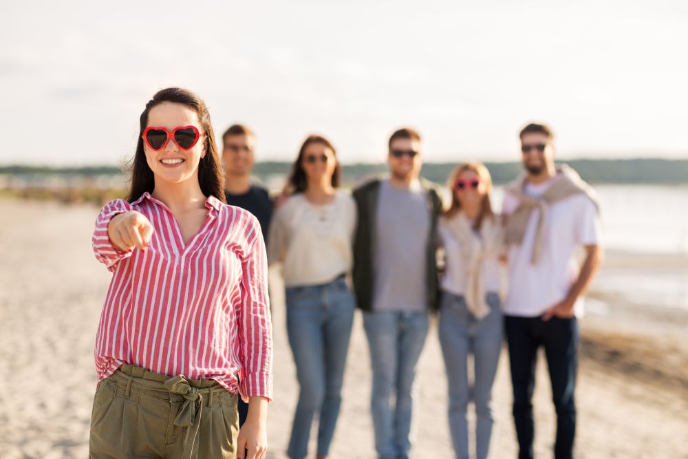 friendship, valentine&rsquo;s day and people concept - happy woman in heart-shaped sunglasses with group of friends on beach in summer pointing to you. woman with friends on beach pointing to you