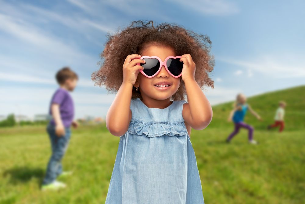 childhood, summer accessory and valentines day concept - happy little african american girl in heart shaped sunglasses over blue sky and meadow background. little african girl in heart shaped sunglasses