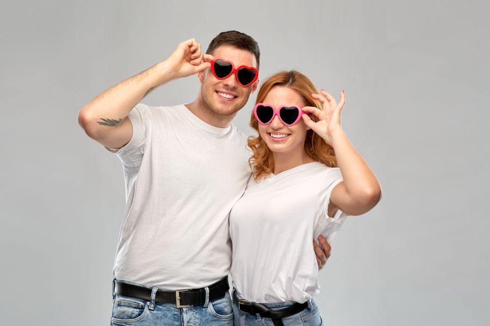 summer accessory, valentines day and love concept - portrait of happy couple in white t-shirts and heart shaped sunglasses over grey background. happy couple in white t-shirts and sunglasses