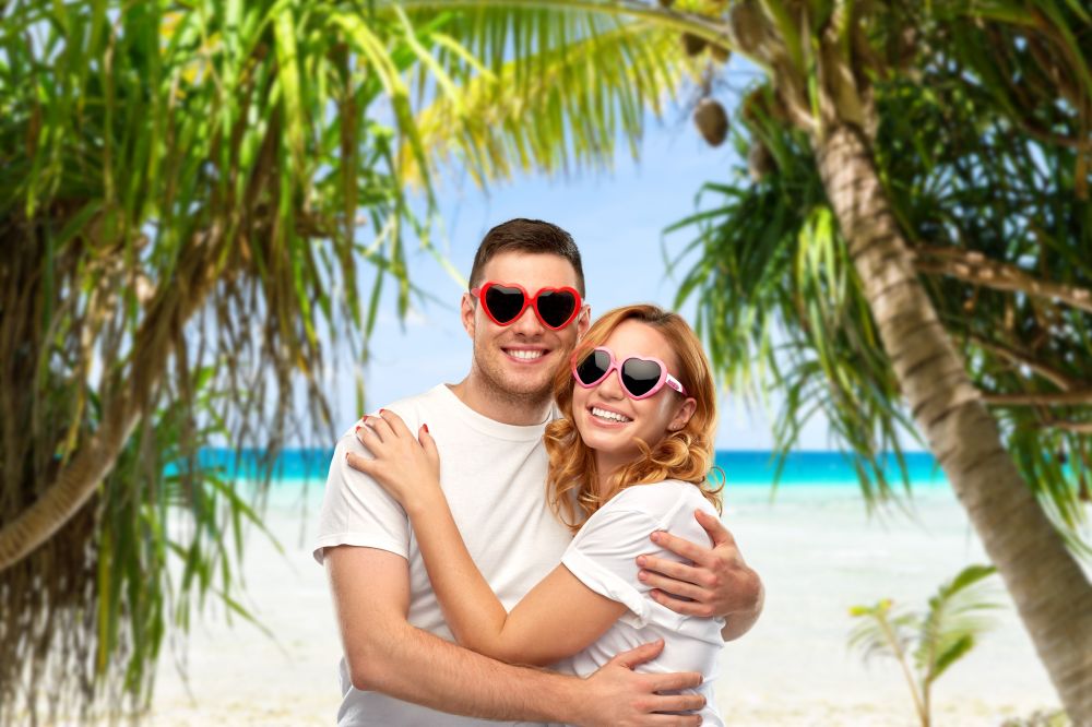 summer accessory, valentine&rsquo;s day and love concept - portrait of happy couple in white t-shirts and heart shaped sunglasses over tropical beach background in french polynesia. happy couple in white t-shirts and sunglasses