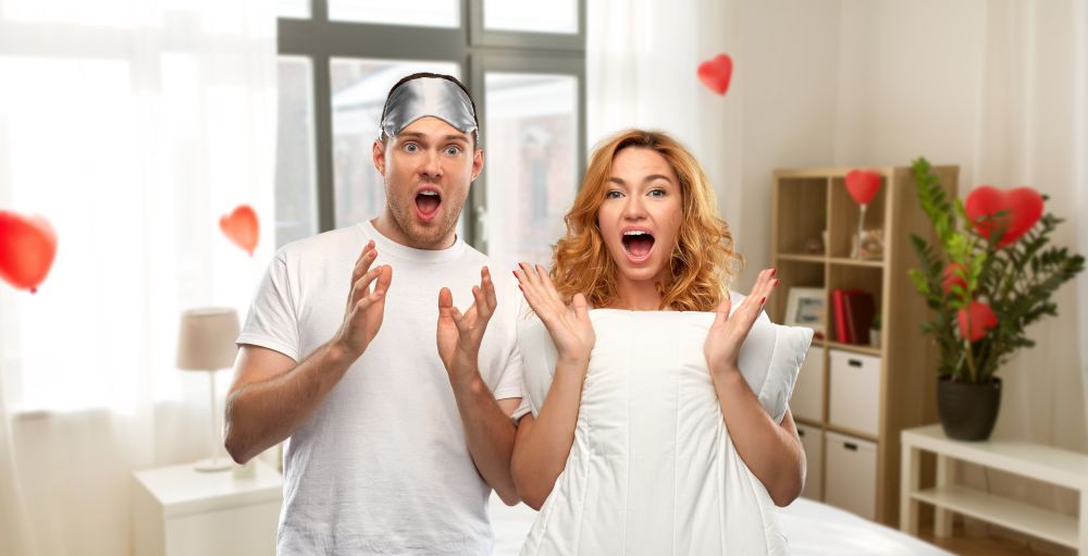 valentine&rsquo;s day and people concept - scared couple in white t-shirts with eye sleeping mask over home bedroom decorated with heart shaped balloons background. scared couple with eye sleeping mask and pillow