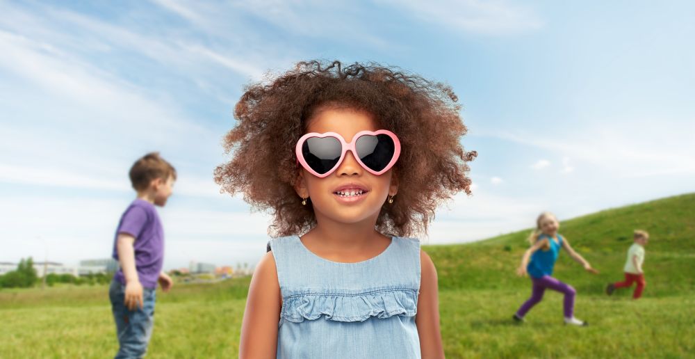 childhood and summer concept - happy little african american girl in heart shaped sunglasses over children playing on meadow background. little african girl in sunglasses at summer