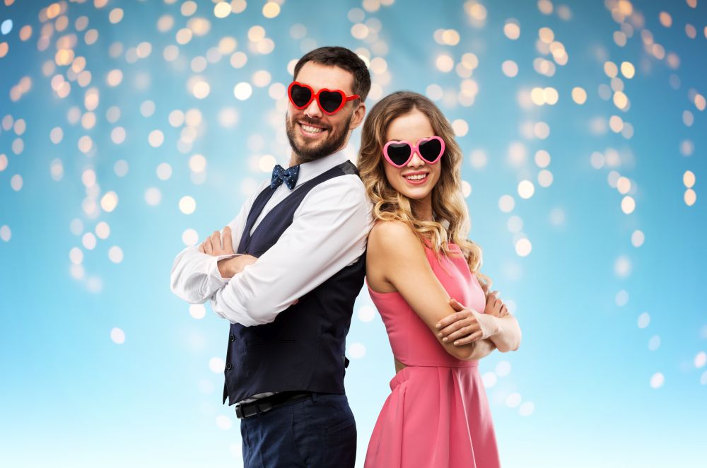 valentines day, love and people concept - happy couple in heart-shaped sunglasses over holiday lights on blue background. happy couple in heart-shaped sunglasses