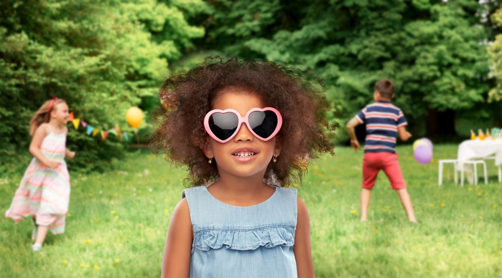 childhood and summer concept - happy little african american girl in heart shaped sunglasses over birthday party at park background. little african girl in sunglasses at summer party